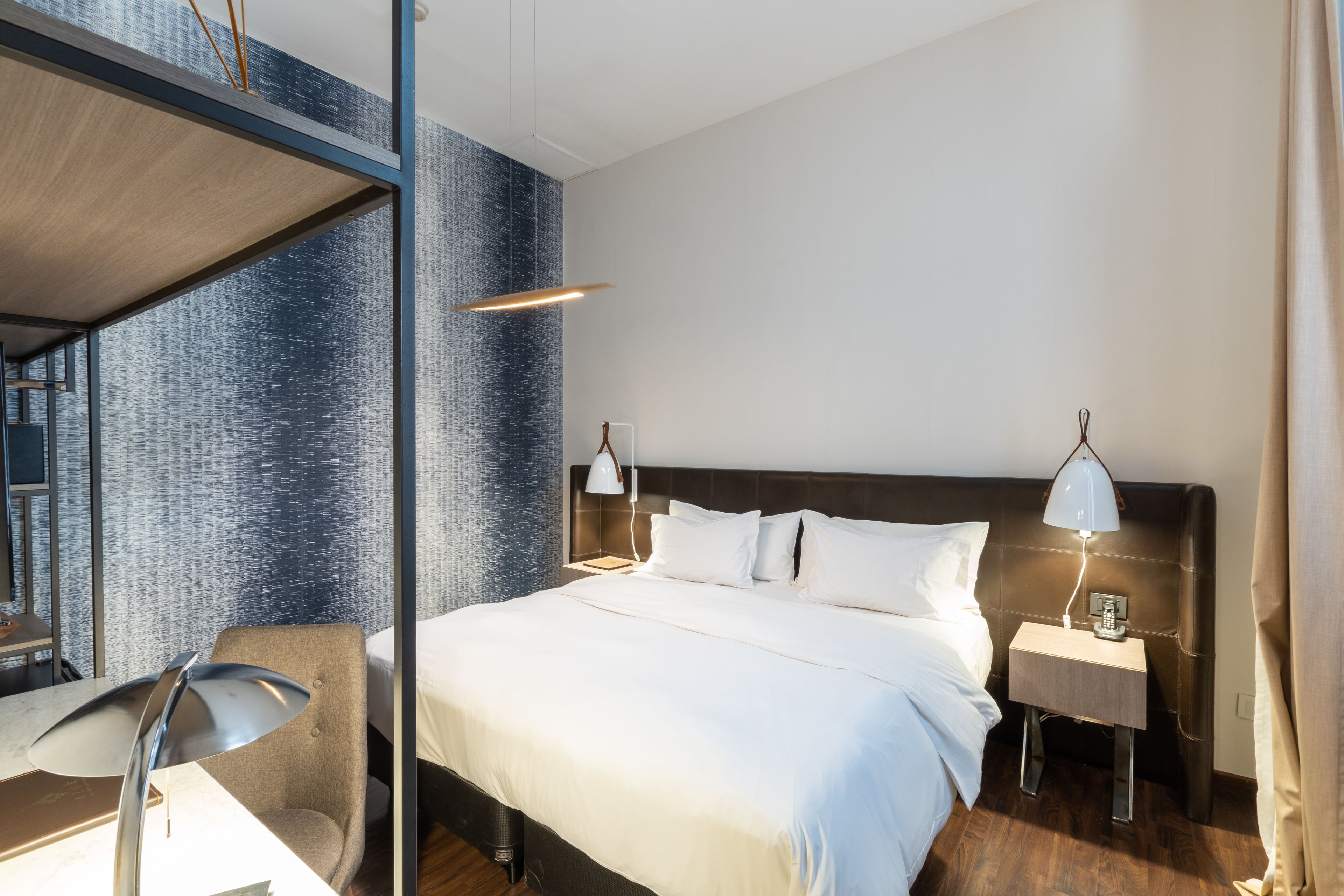 Una habitación de hotel moderna con una cama grande, dos mesitas de noche con lámparas, una silla y vistas a la ciudad a través de un muro de cristal.