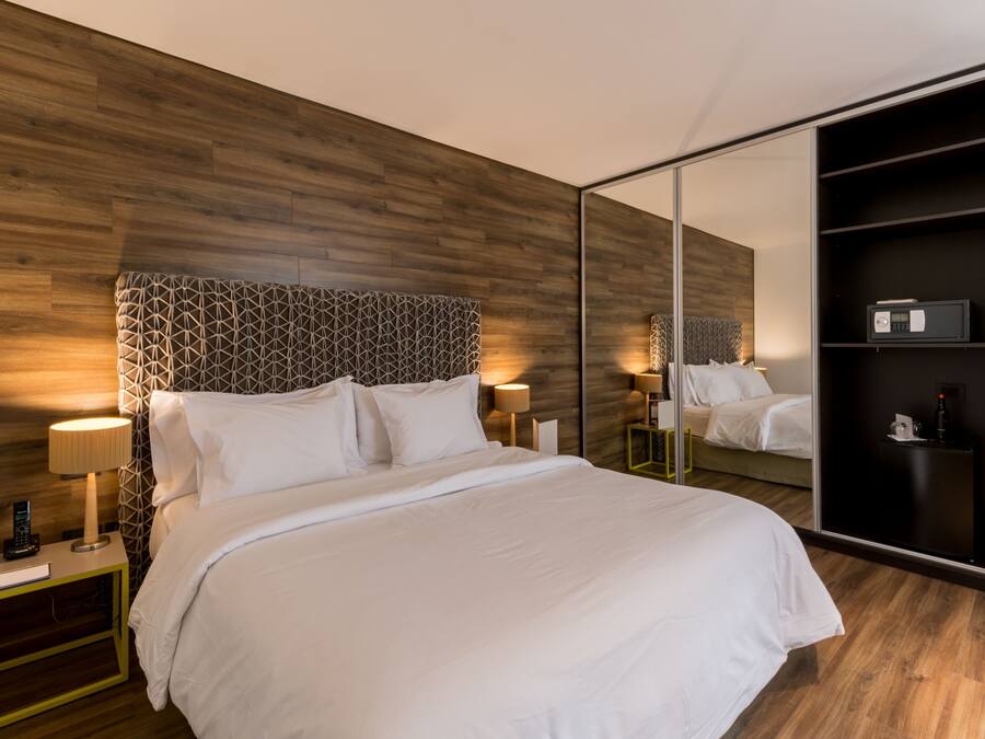 Una habitación de hotel moderna con cabecera de madera, un espejo grande, una mesita de noche con lámpara y un armario.