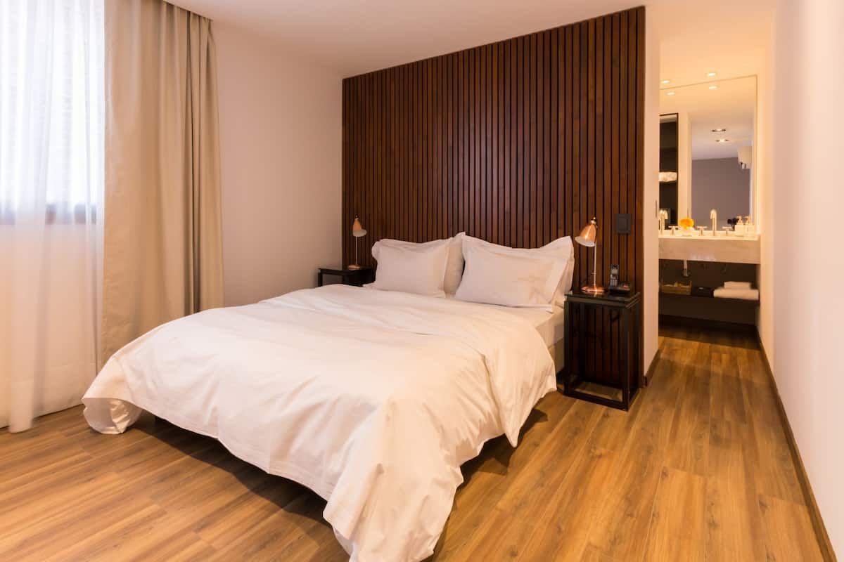 A modern hotel room with a large bed, wooden panel wall, and a bathroom with a mirror and sink.