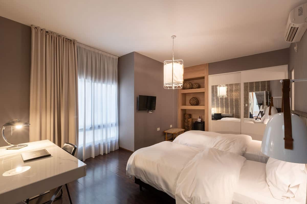 A hotel room with a bed, a desk with a lamp, a TV, a mirror, and a window with curtains.