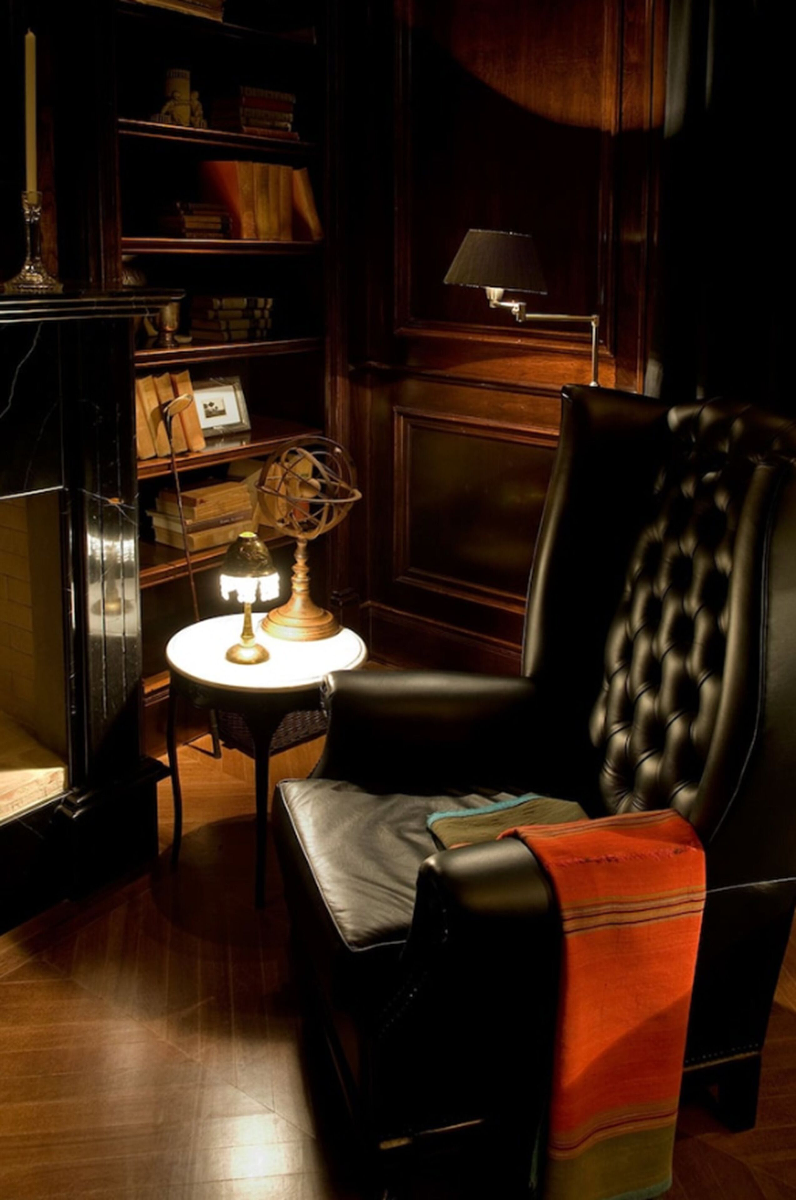 A dark room with a leather armchair, a small round table with a lamp, and a bookshelf with various items.