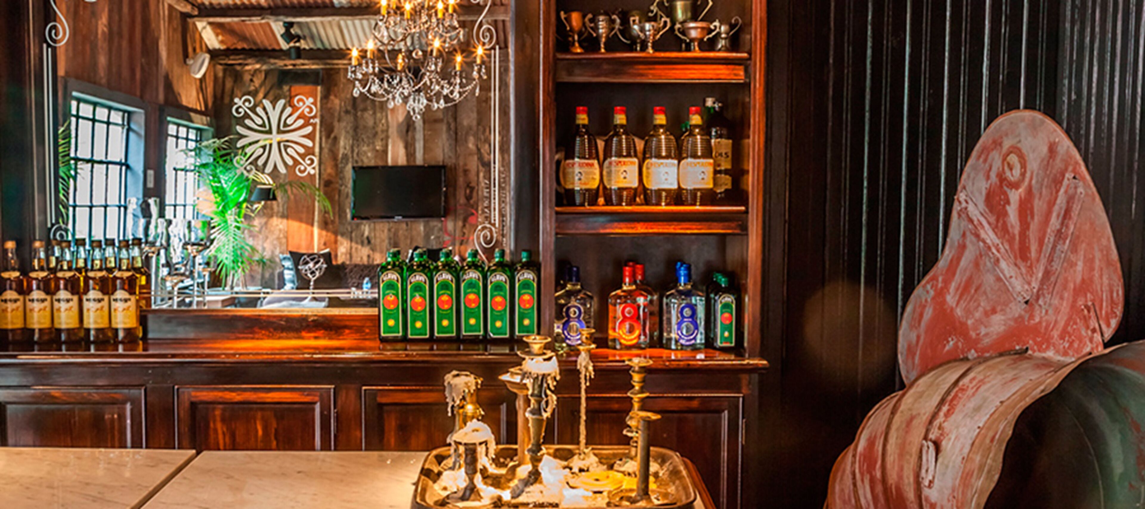 A bar area with a wooden bar counter, shelves with bottles, a television, and a chandelier.