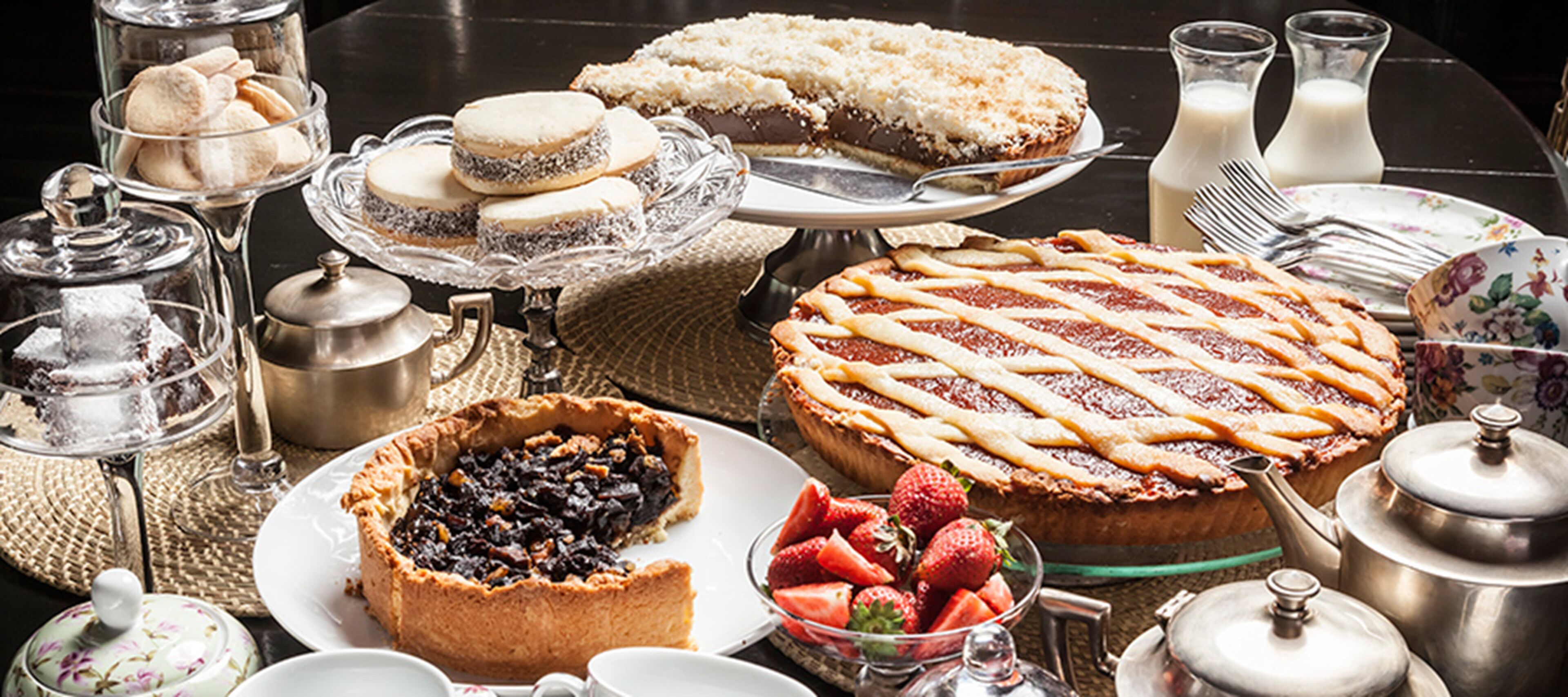 A variety of desserts including pies, tarts, and cookies on a table with tea sets and milk bottles.