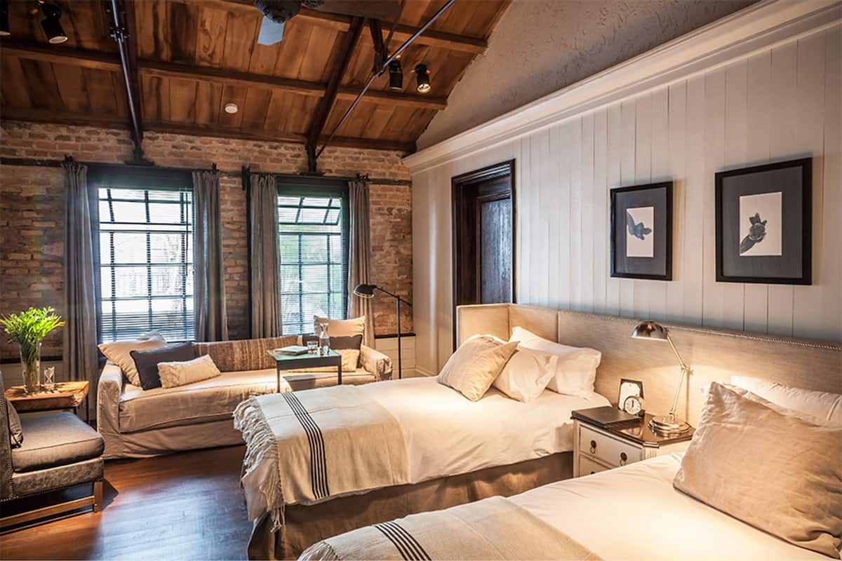 A hotel room with a rustic wooden ceiling, two beds with white linens, a nightstand with a lamp, and framed artwork on the wall.