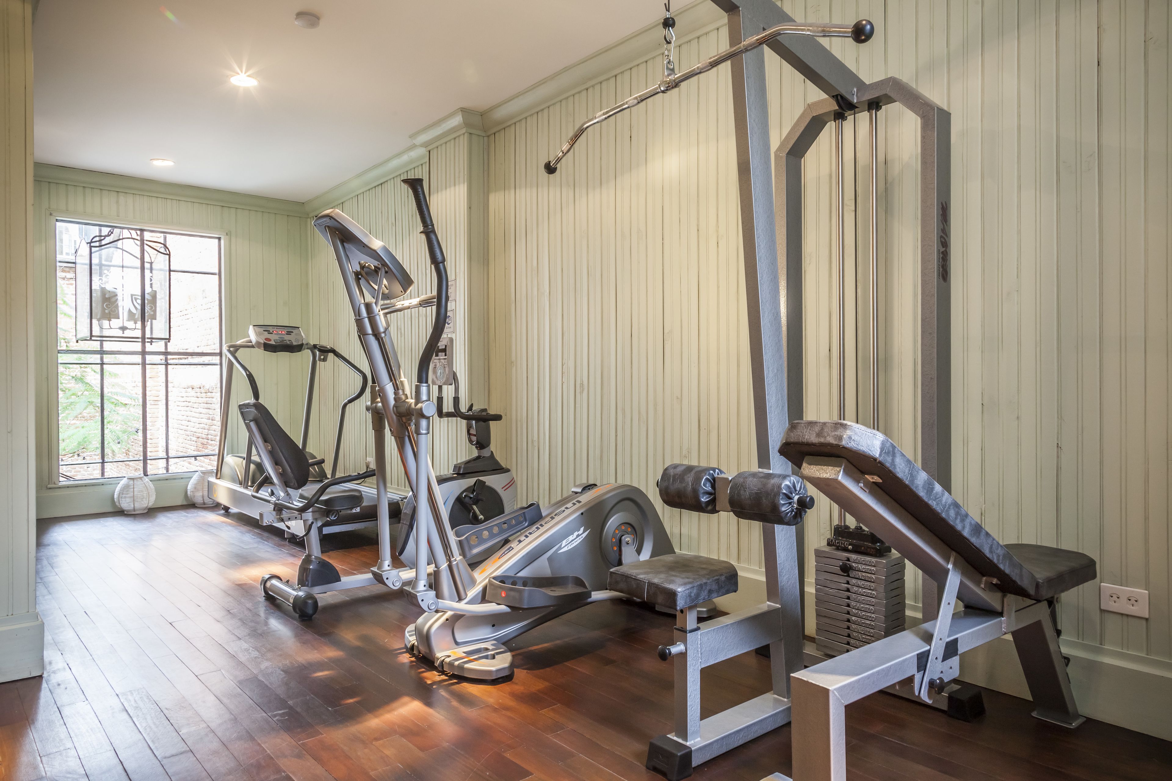 A gym with various exercise equipment, including treadmills and weight machines, in a room with wooden floors and a large window.