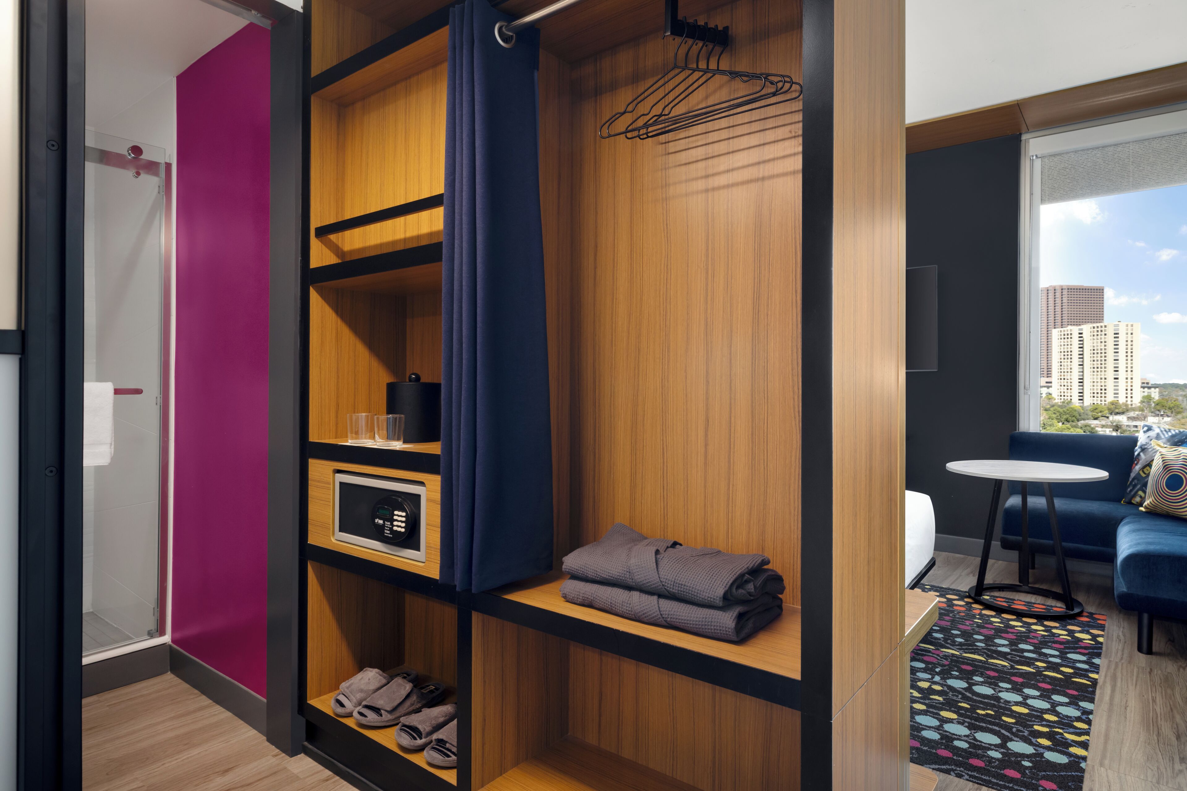 A modern hotel room with a wooden wardrobe, a small table, a blue sofa, and a city view.