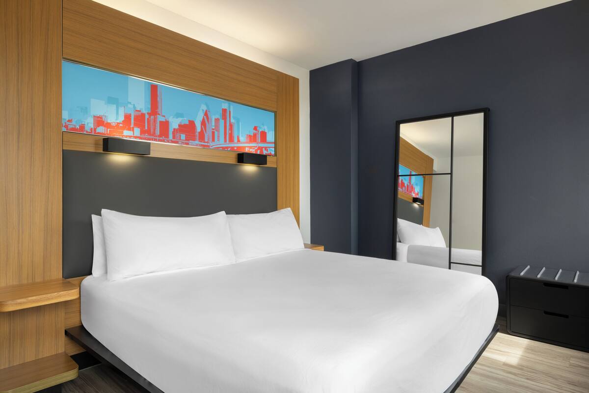 Una habitación de hotel moderna con una cama grande, una obra de arte con un paisaje urbano, un espejo y una mesita de noche.