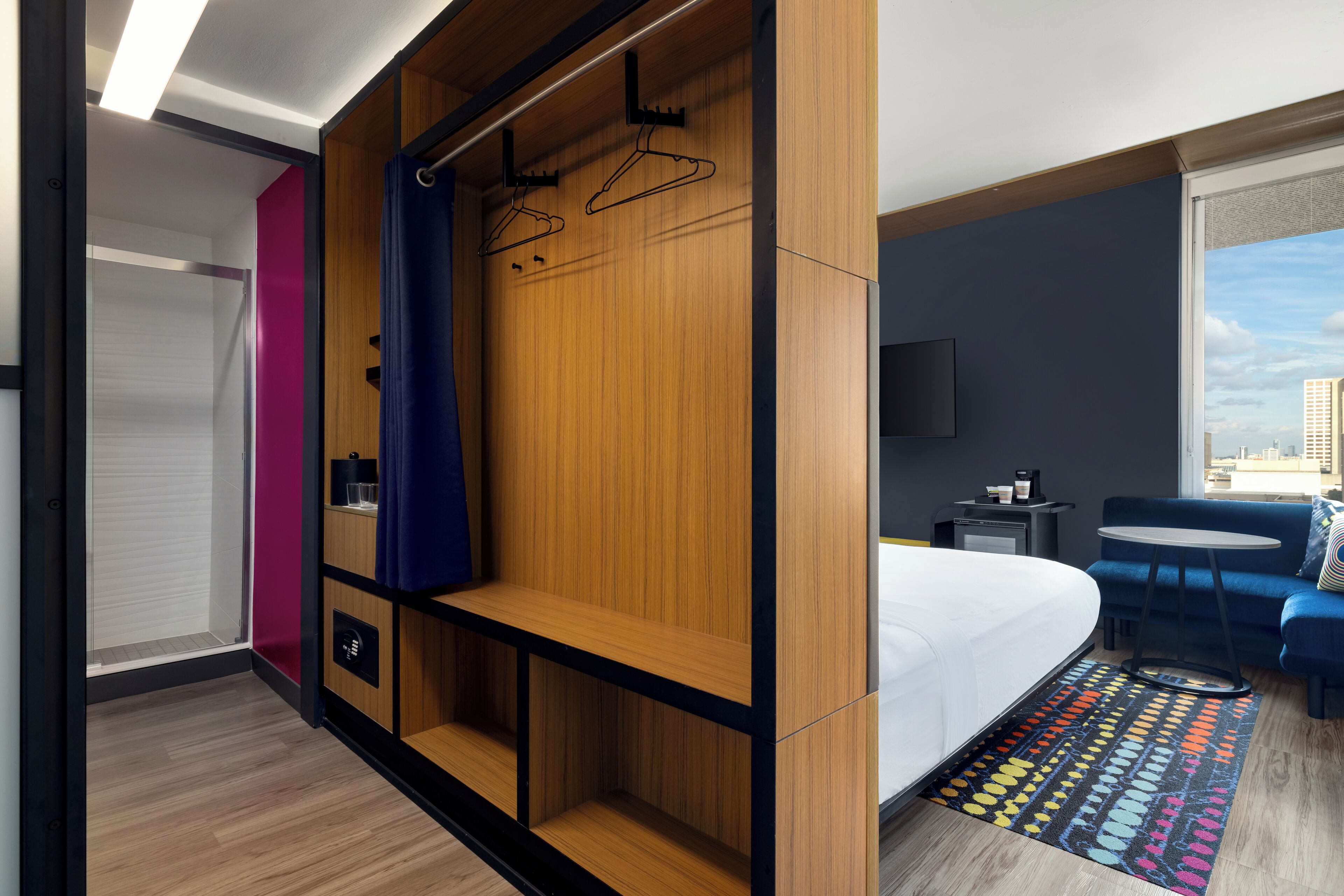 A hotel room with a bed, a seating area, a wardrobe with hangers, a television, and a city view.