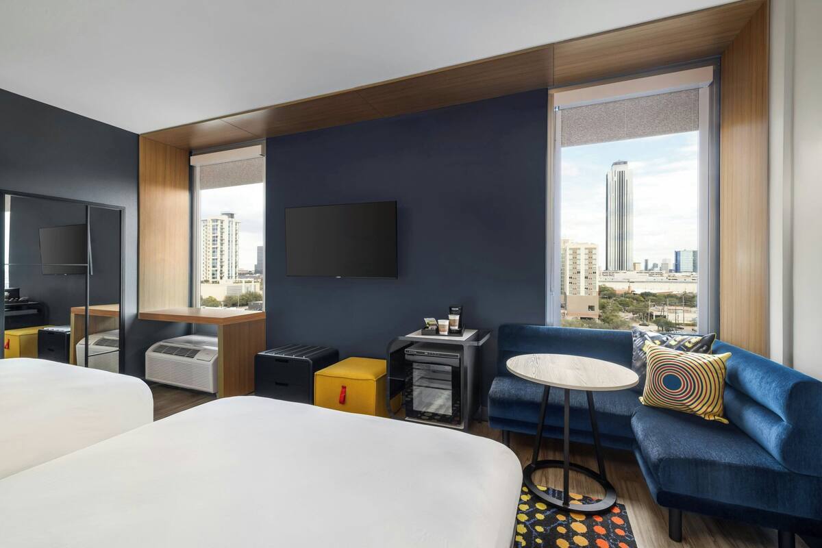 Una habitación de hotel moderna con una cama, un sofá, una mesita, un televisor y una ventana con vistas a la ciudad.