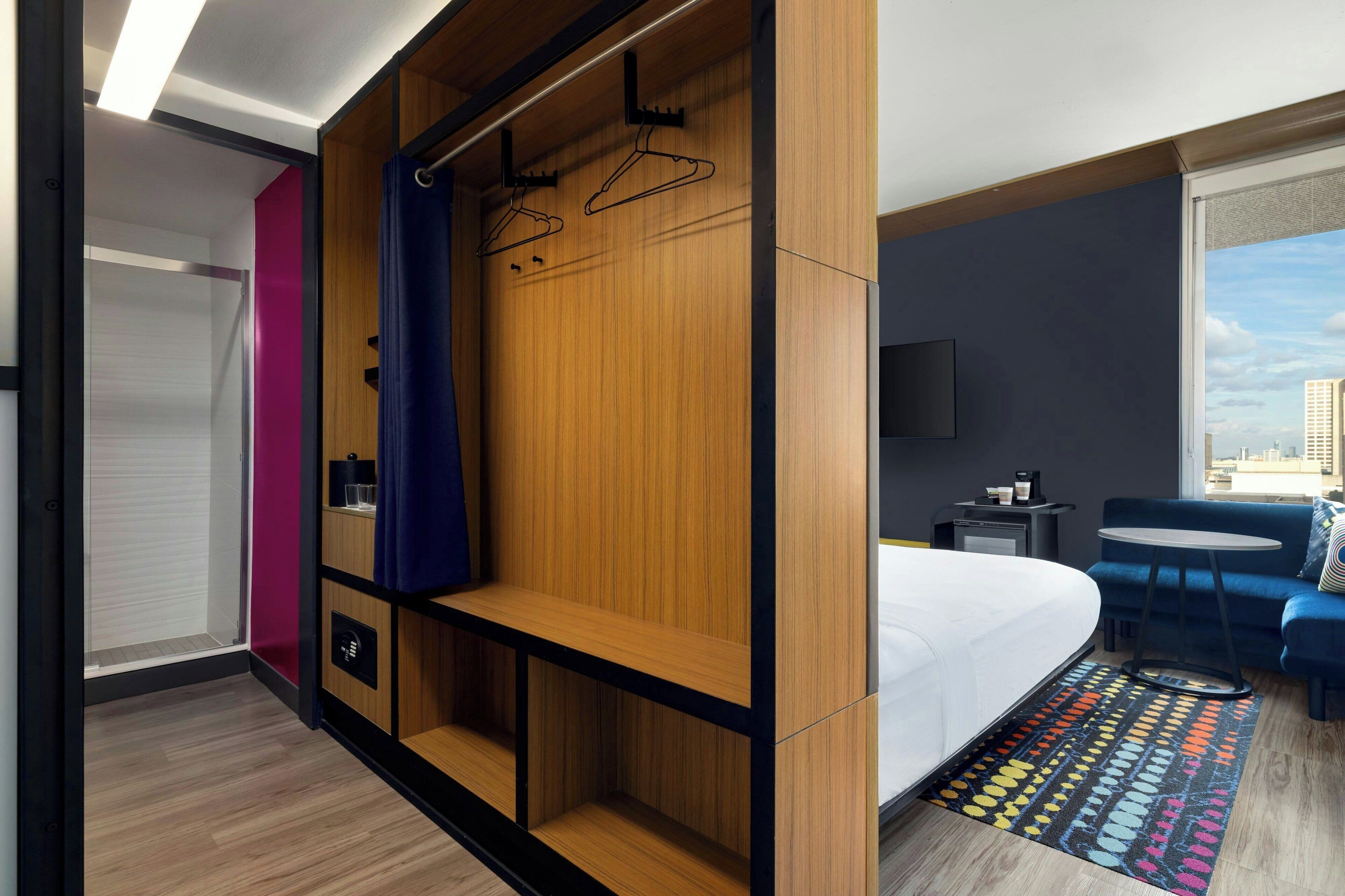 A modern hotel room with a large bed, a seating area with a table, a TV, and a wardrobe with hangers.