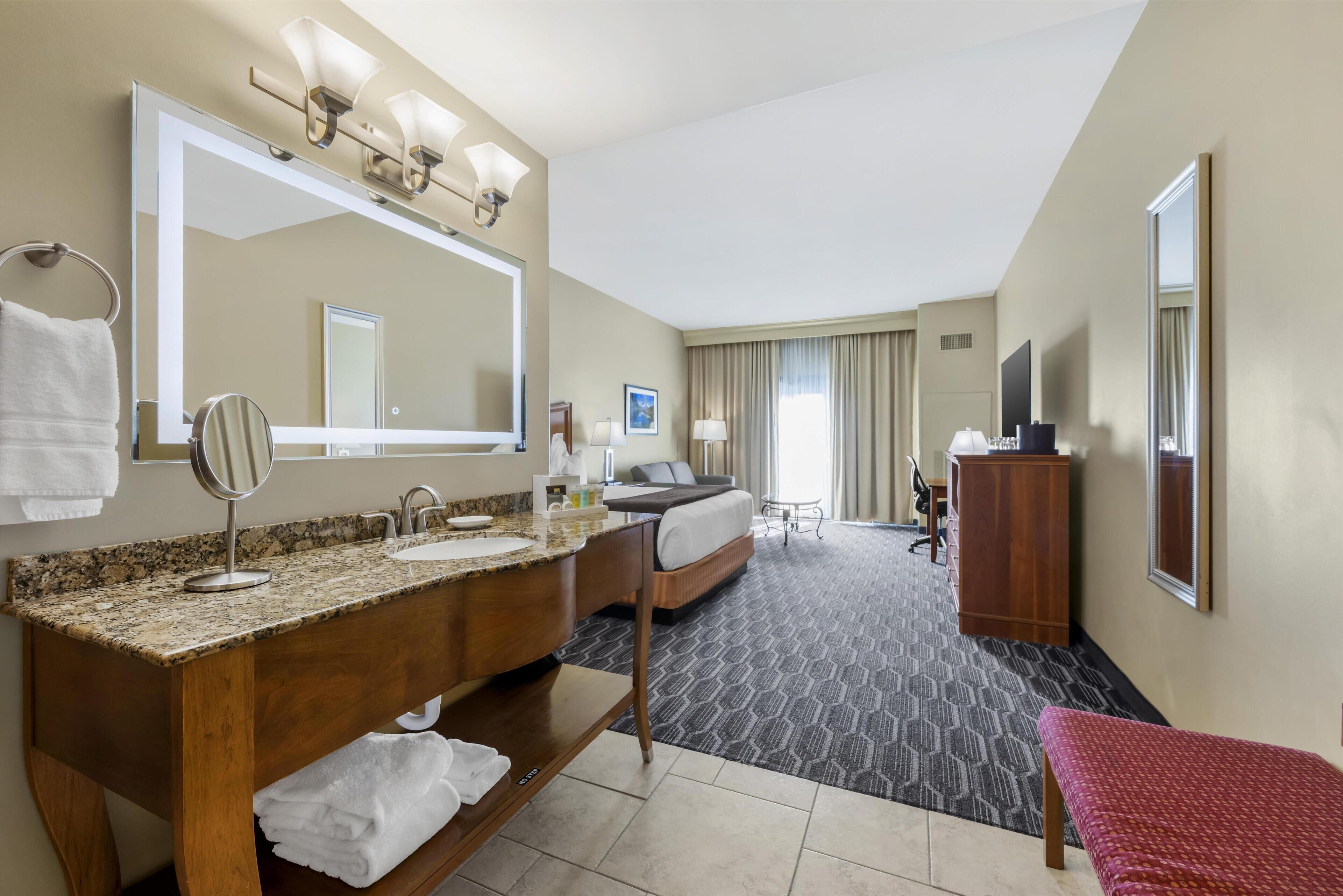 A hotel room with a large mirror, double sink vanity, bed, bedside table, and a red bench.
