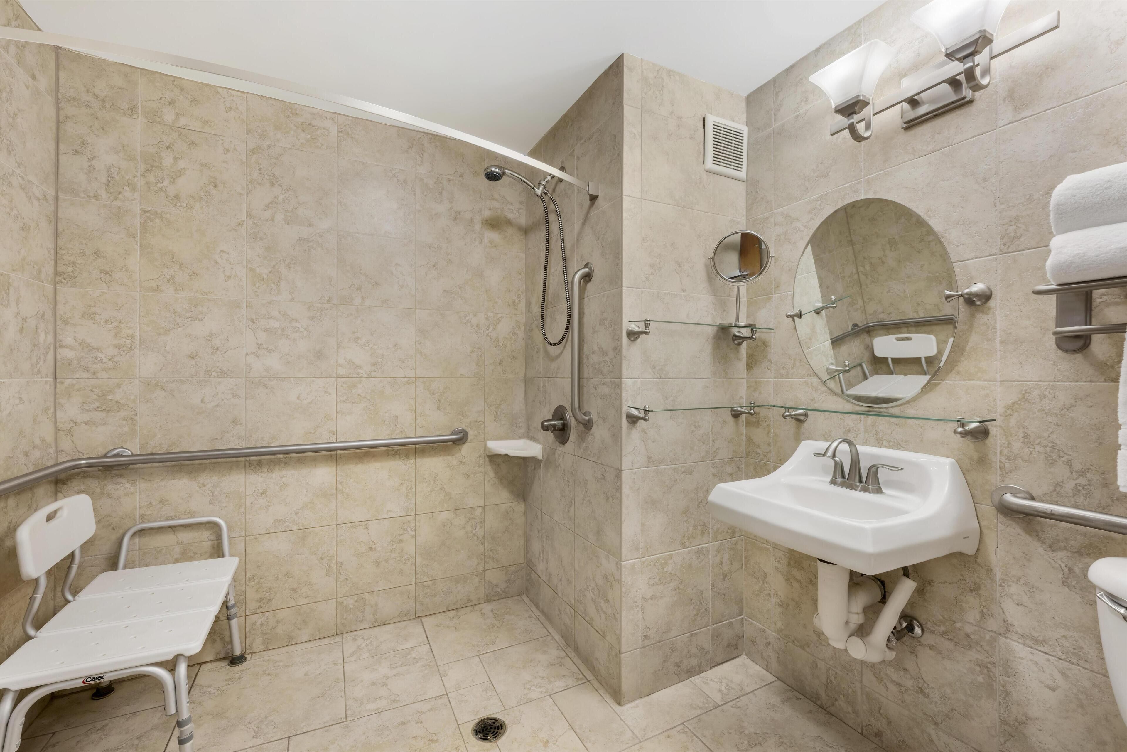 A bathroom with a shower, a sink, a toilet, a bench, and a handrail.