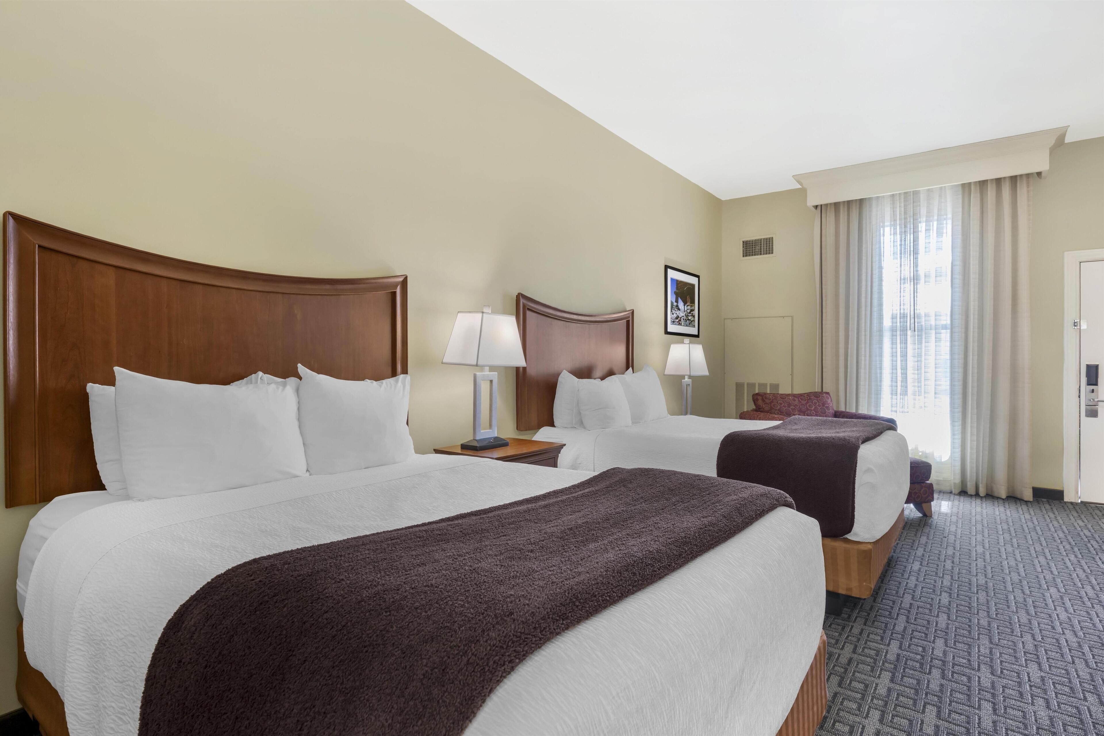 Habitación de hotel con dos camas, un ventanal con cortinas y suelo alfombrado.