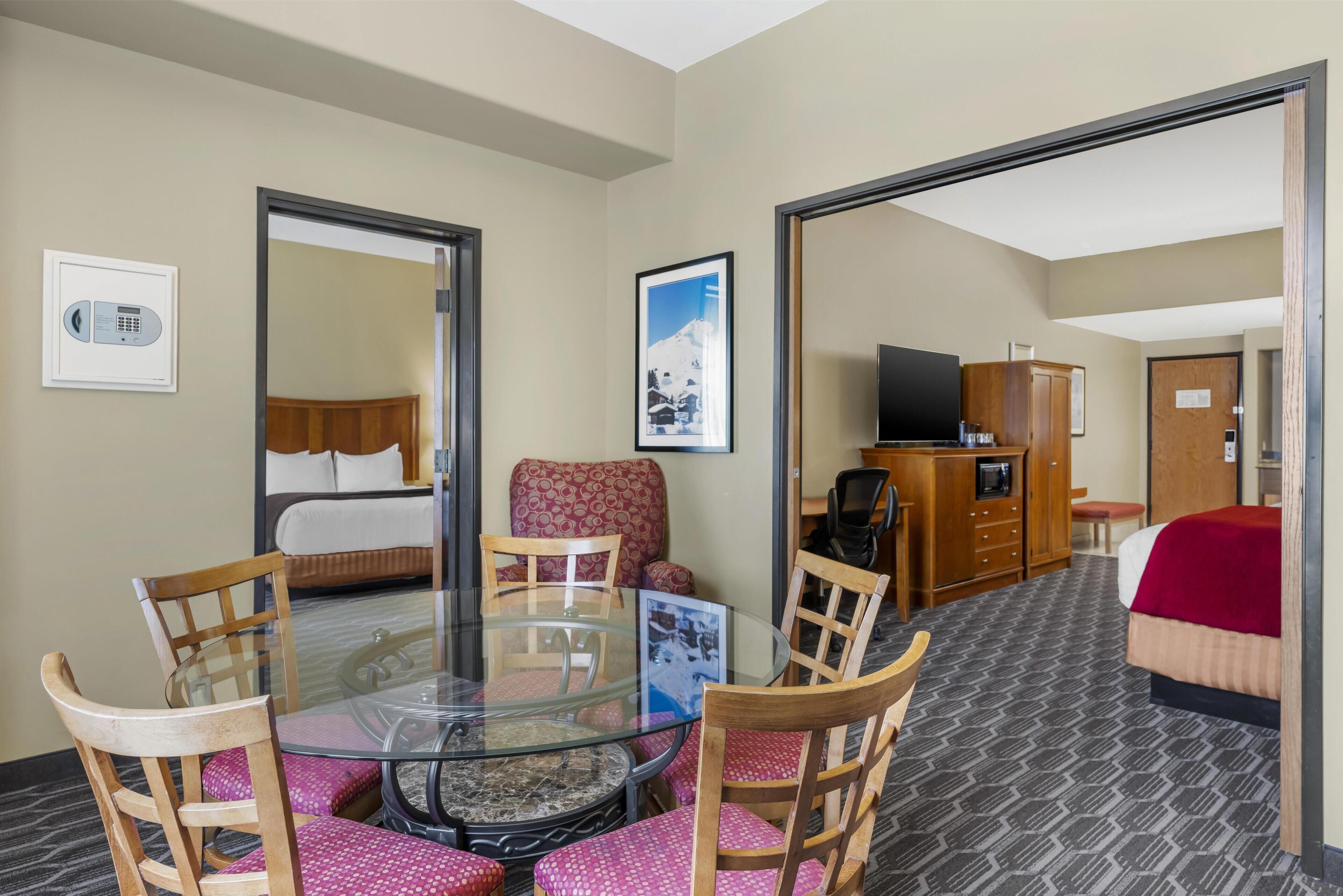 A hotel room with a glass dining table, wooden chairs, a bed with a red blanket, a TV, and a wardrobe.