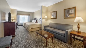 A hotel room with a bed, sofa, coffee table, desk, lamp, and artwork on the walls.