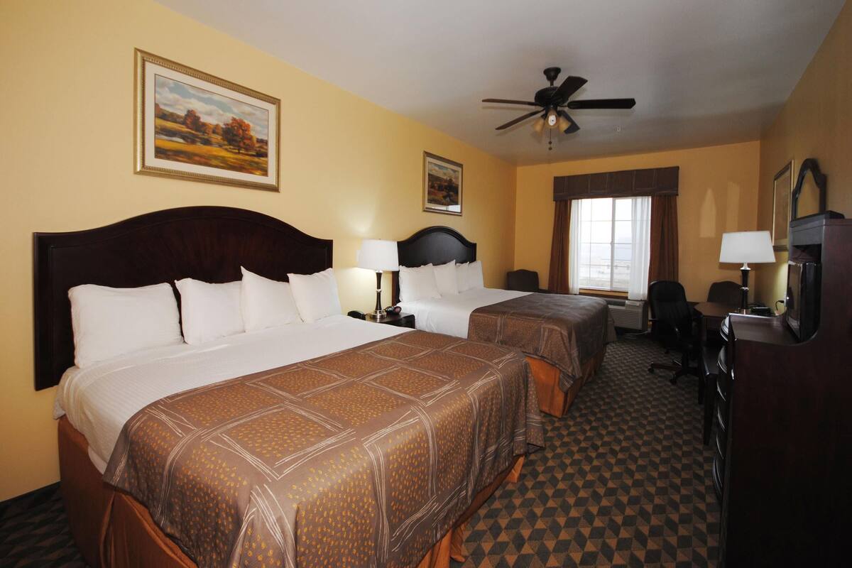 A hotel room with two beds, a ceiling fan, a desk with a chair, a TV, and a window with curtains.