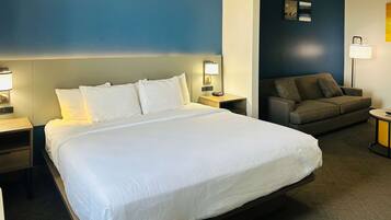 A hotel room with a large bed, a sofa, a nightstand with a lamp, and a wall-mounted artwork.