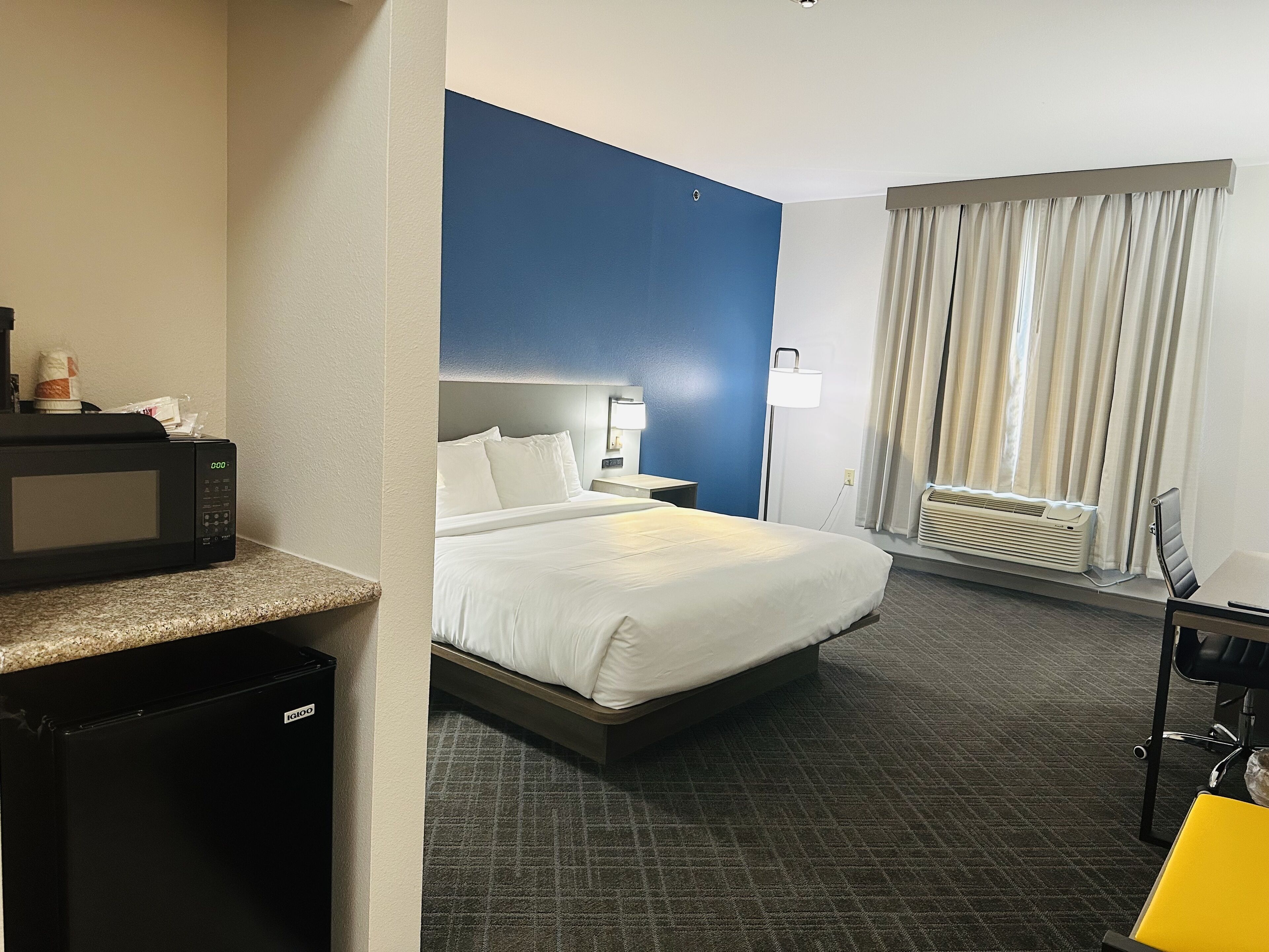 A hotel room with a bed, a desk, a microwave, a mini-fridge, a lamp, a window with curtains, and an air conditioning unit.