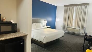 A hotel room with a bed, a desk, a microwave, a mini-fridge, a lamp, a window with curtains, and an air conditioning unit.