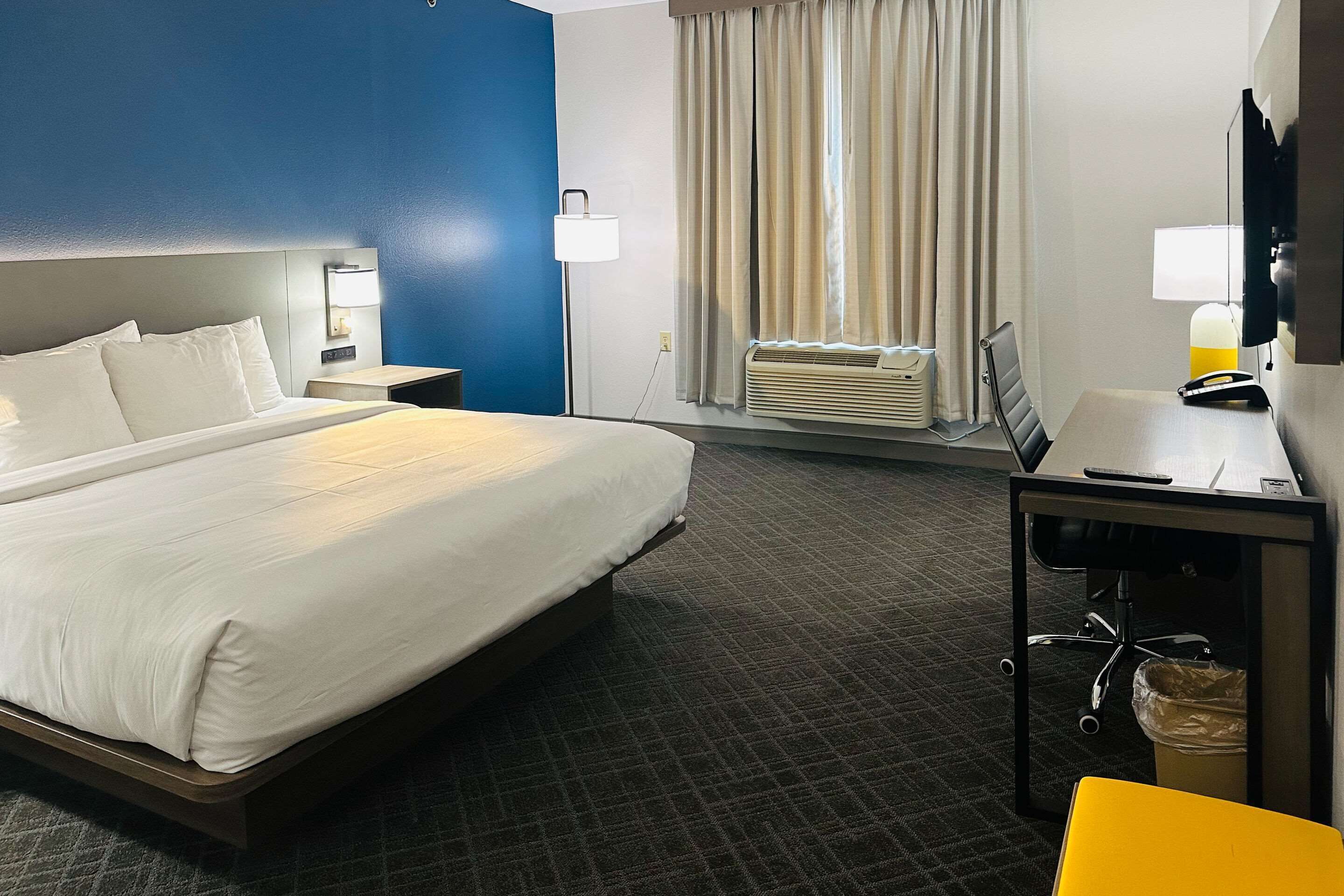 Habitación de hotel con una cama grande, un escritorio con silla, una lámpara de escritorio, un televisor y una ventana con cortinas.