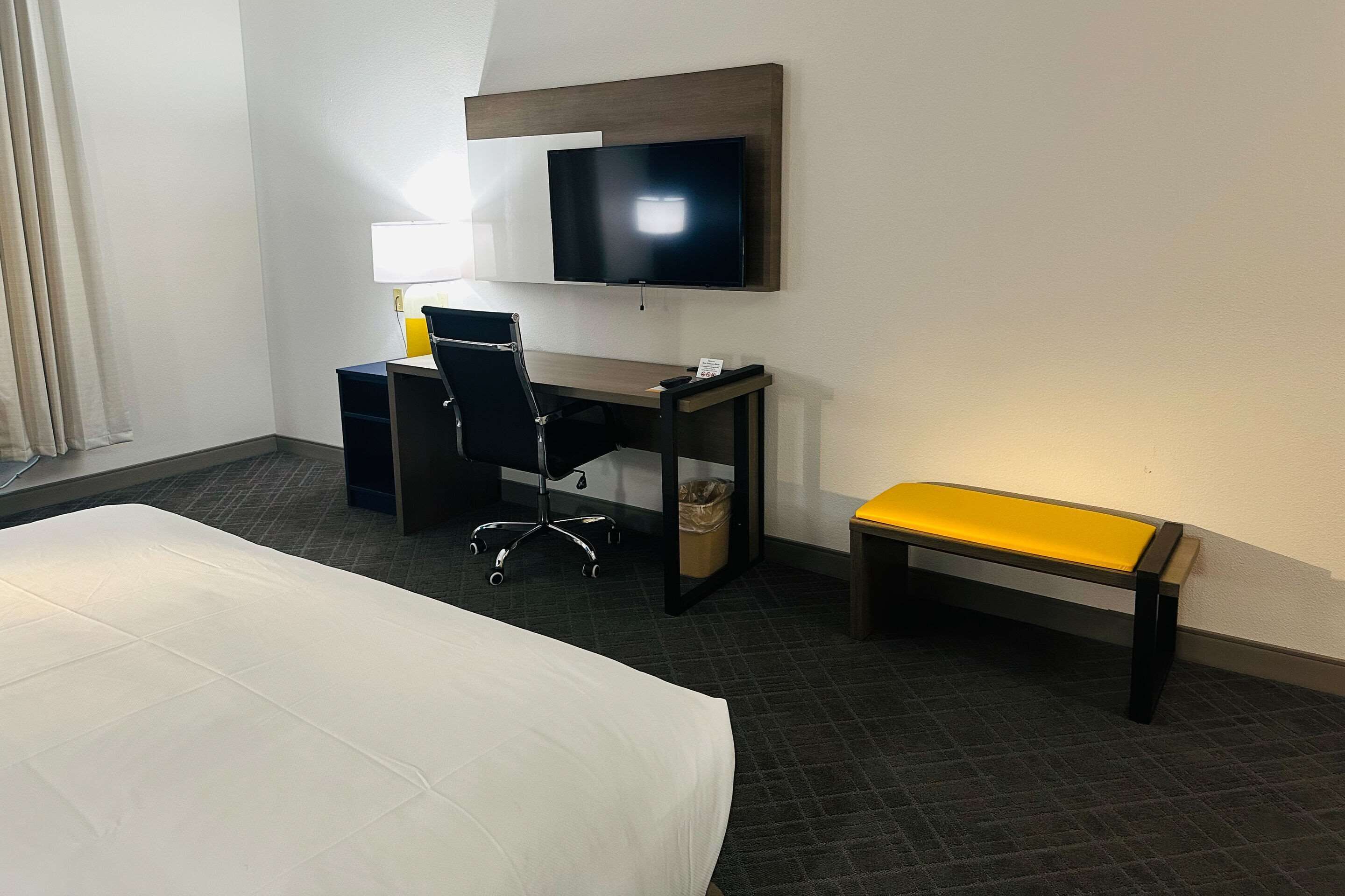 A hotel room with a bed, a desk with a chair, a TV mounted on the wall, and a bench.