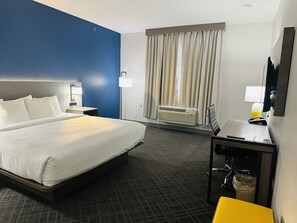 Une chambre d’hôtel avec un grand lit, un bureau avec une chaise, une lampe de bureau, un téléviseur fixé au mur et une fenêtre avec des rideaux.