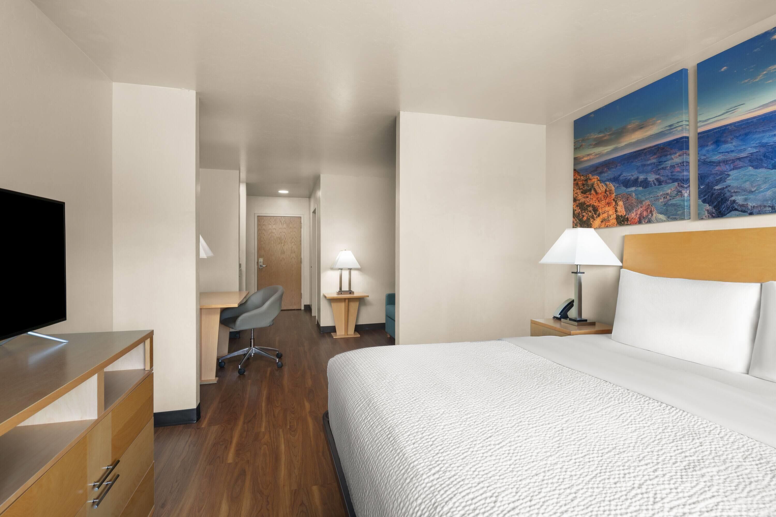A hotel room with a bed, a desk, a chair, a TV, and a painting on the wall.