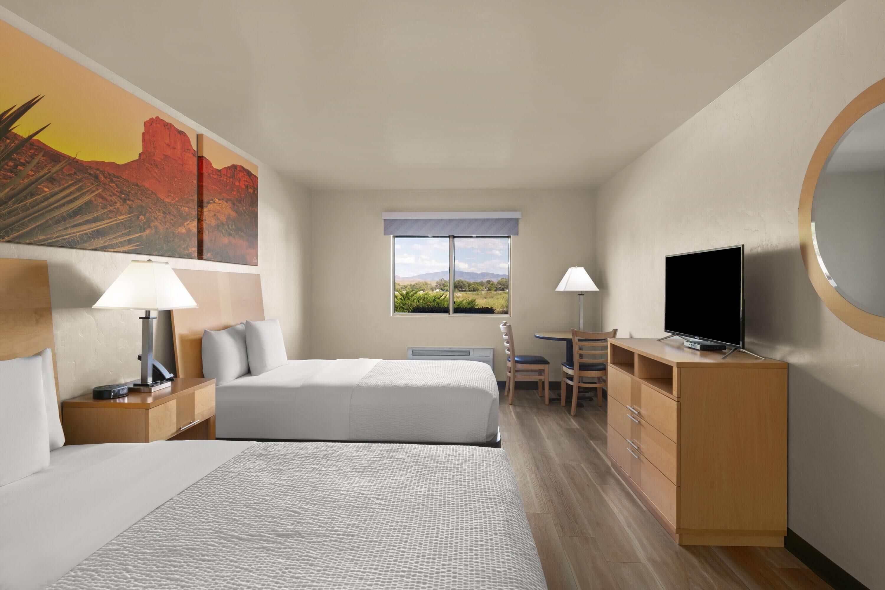 A hotel room with a bed, bedside tables, a desk, a chair, a television, and a large window with a view of the outdoors.