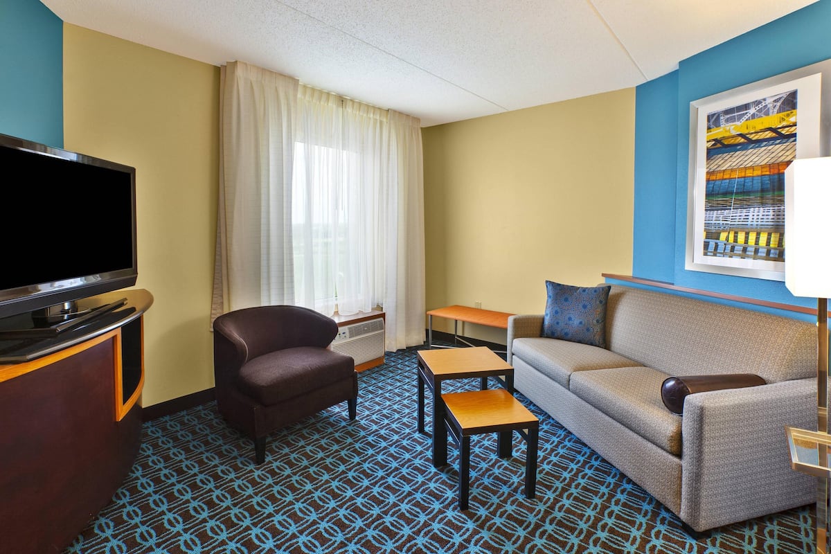 A hotel room with a flat-screen TV, a sofa, two armchairs, a small table, a window with curtains, and a framed picture on the wall.