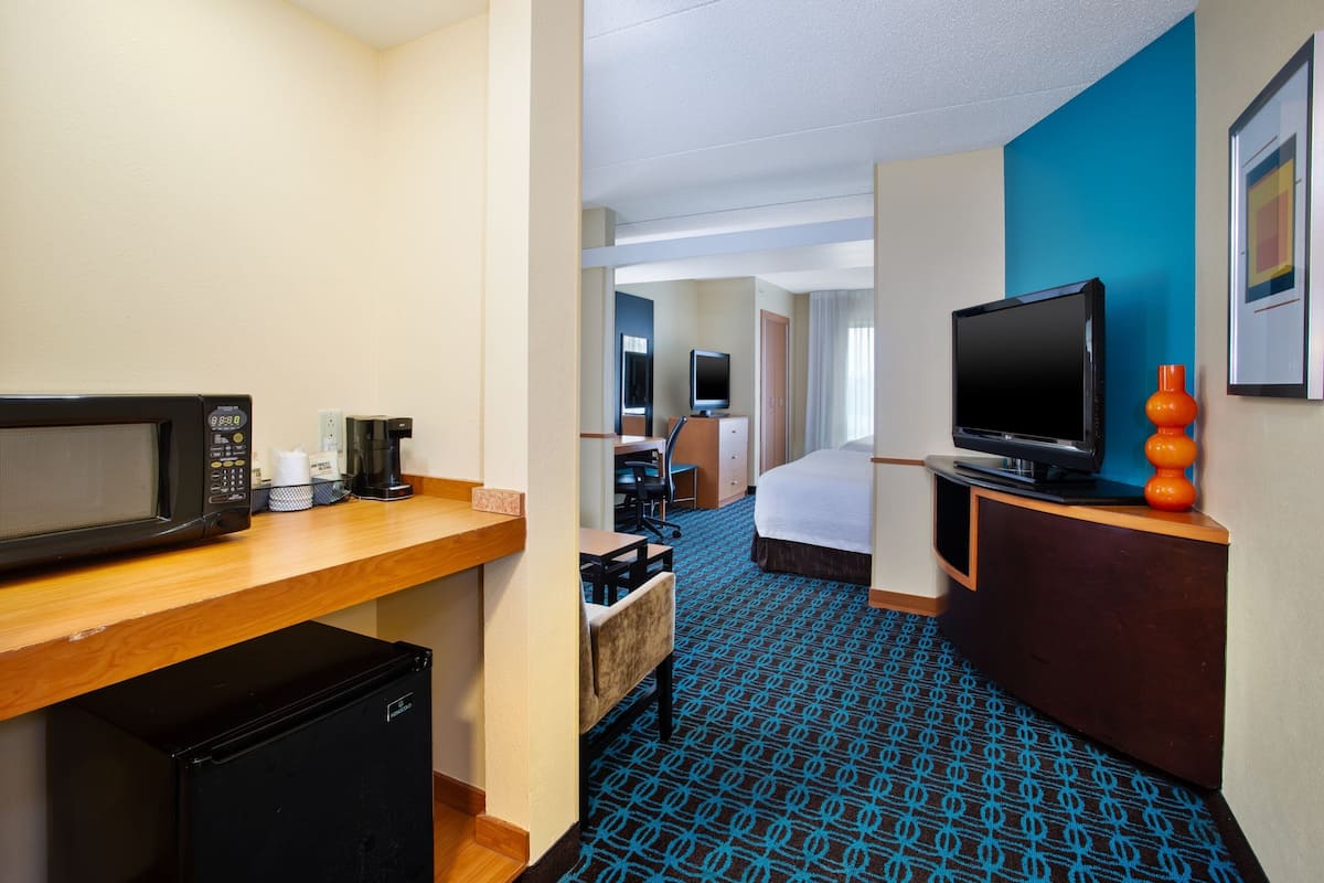 A hotel room with a kitchenette, a bed, a desk, a television, and a blue patterned carpet.