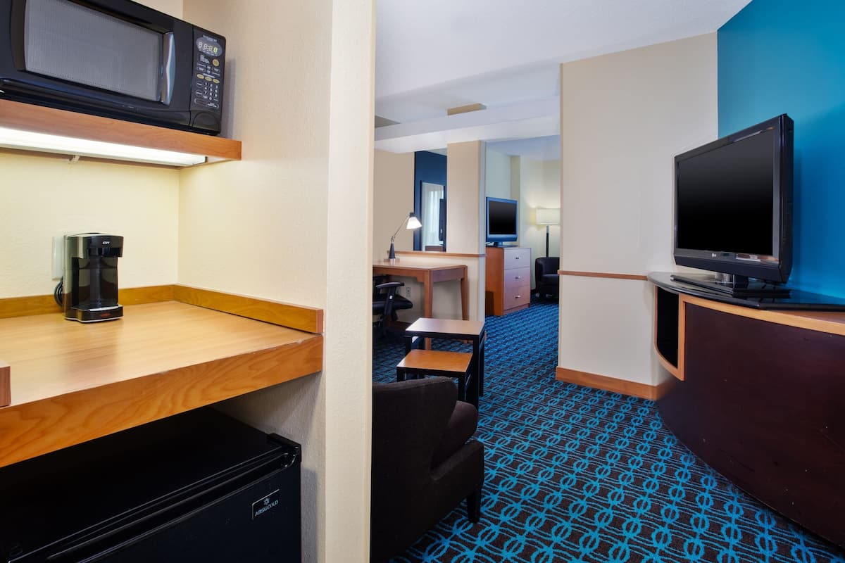 A hotel room with a microwave, coffee maker, TV, desk, chair, and a patterned carpet.