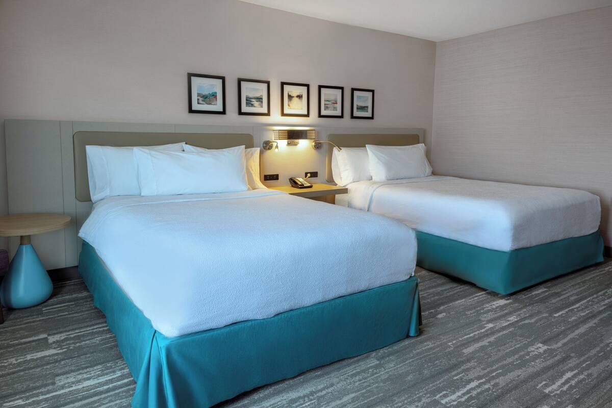A hotel room with two beds, a nightstand, a lamp, and framed pictures on the wall.