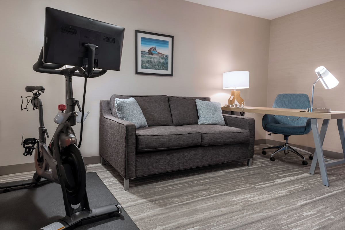 A room with a grey sofa, a desk with a chair, a standing exercise bike, a TV mounted on the wall, and a framed picture.