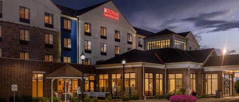 A multi-story hotel with a prominent Hilton Garden Inn sign, a covered outdoor seating area, and a parking lot.