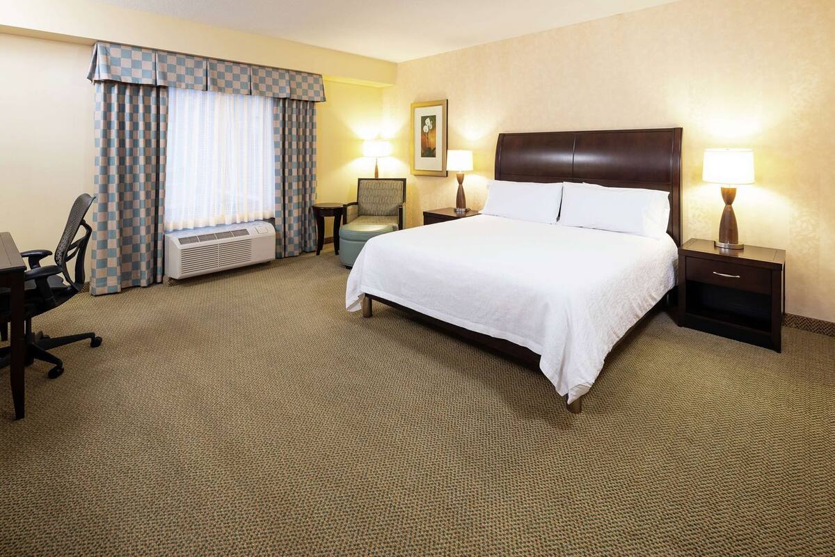 A hotel room with a bed, two bedside tables with lamps, a desk with a chair, a window with curtains, and a radiator.