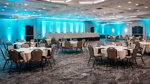 A large conference room with multiple round tables and chairs, a stage with curtains, and a carpeted floor.
