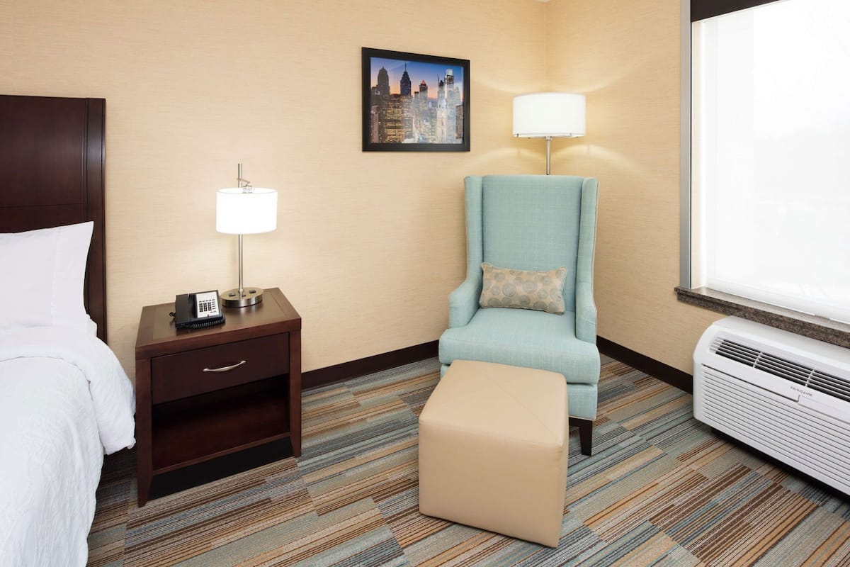 Habitación de hotel con cama, escritorio con teléfono, silla con cojín, lámpara de pie y unidad de aire acondicionado.
