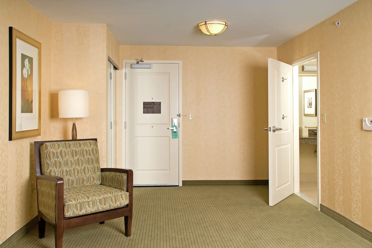 Un pasillo de hotel con un sillón estampado, una puerta cerrada y una puerta abierta que da a un baño.