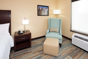Habitación de hotel con cama, escritorio con lámpara, sillón con cojín, reposapiés y ventana grande.