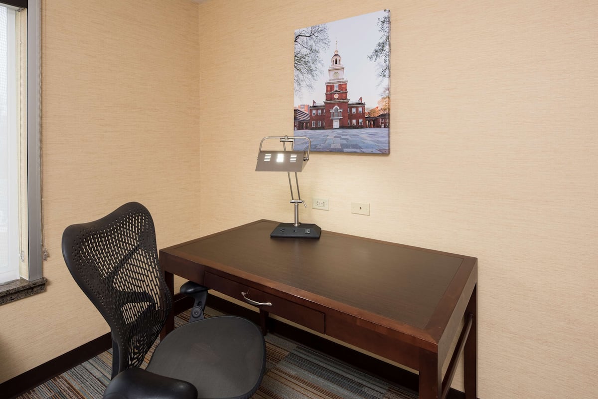 Habitación de hotel con un escritorio de madera, una silla de malla negra, una lámpara de escritorio y un cuadro con una vista de un edificio en la pared.
