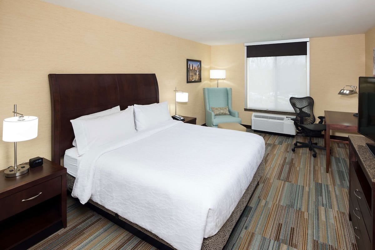 Habitación de hotel con una cama grande, un escritorio con silla, televisión, lámpara y ventana.