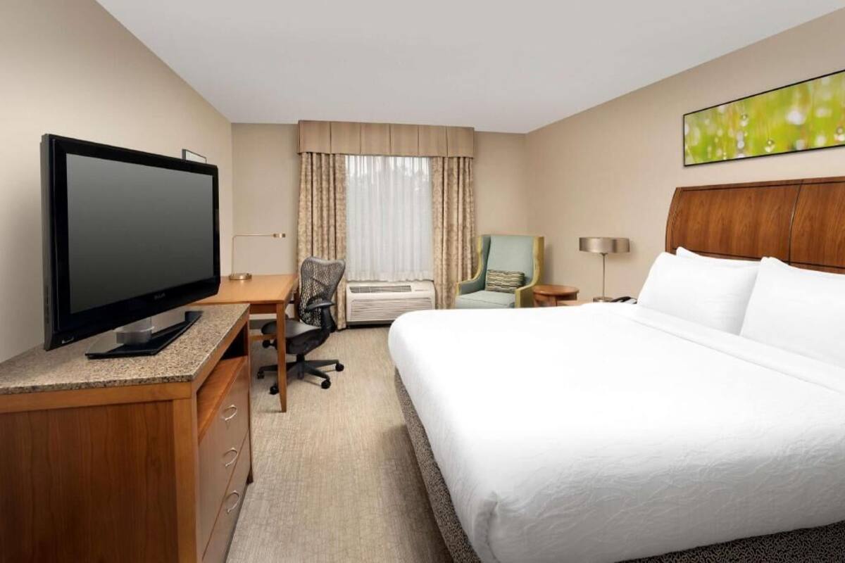 A hotel room with a bed, a desk with a flat-screen TV, an air conditioning unit, a chair, and a lamp.