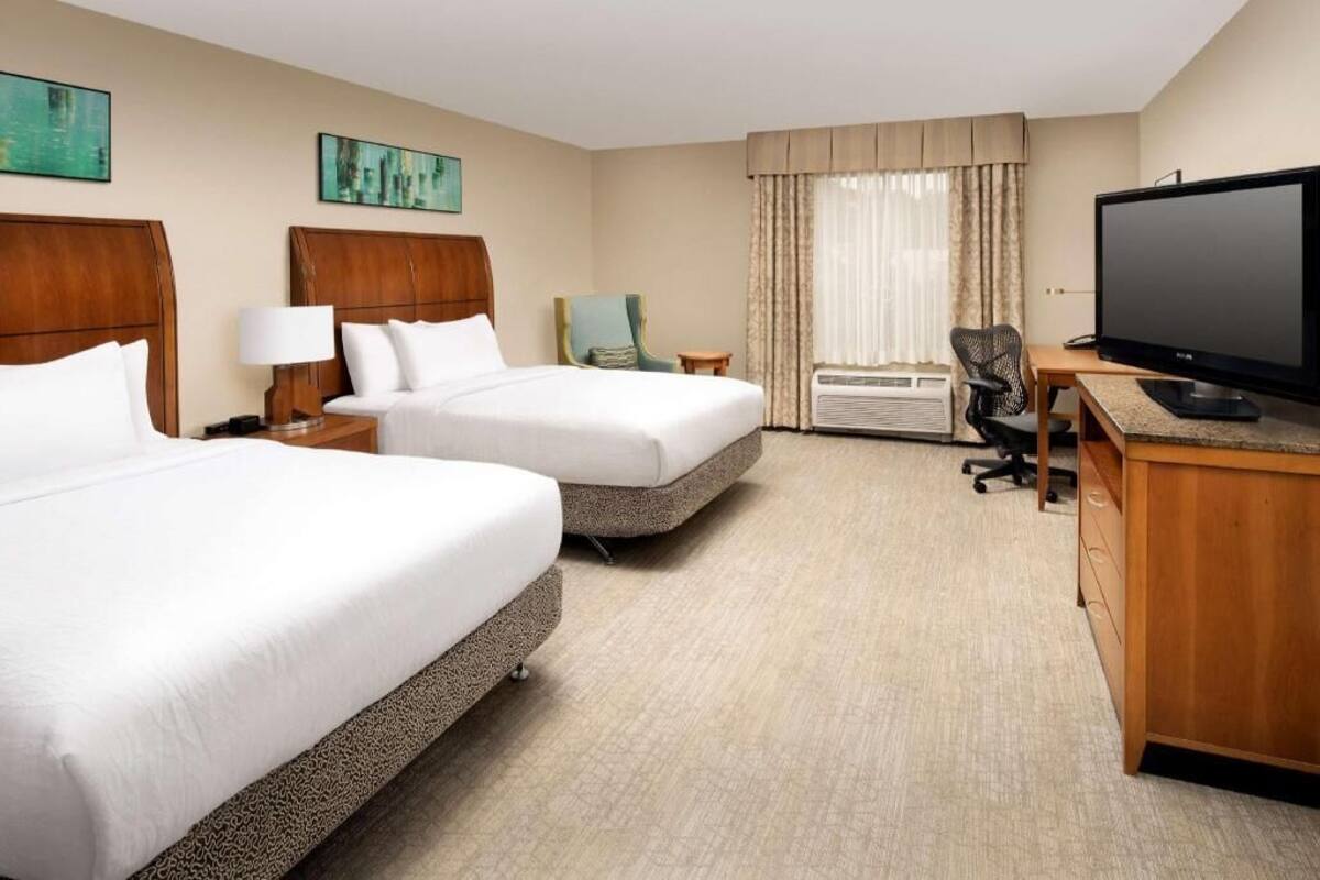A hotel room with two beds, a desk with a television, a chair, an air conditioning unit, and artwork on the walls.