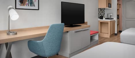 Une chambre d’hôtel moderne avec un bureau en bois, une chaise bleue, un téléviseur à écran plat, une petite kitchenette et un lit.
