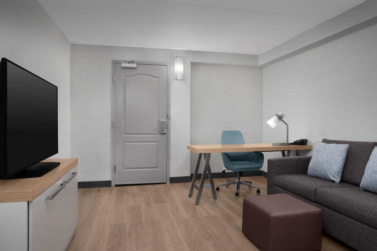 A modern hotel room with a flat-screen TV, a wooden desk, a grey sofa, and a brown ottoman.