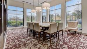 A spacious dining area with a large table and multiple chairs, large windows with a view of the outdoors, and a patterned carpet.