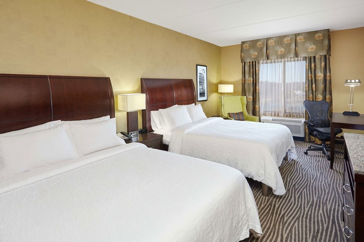 A hotel room with two beds, a desk, a chair, a lamp, and a window with curtains.