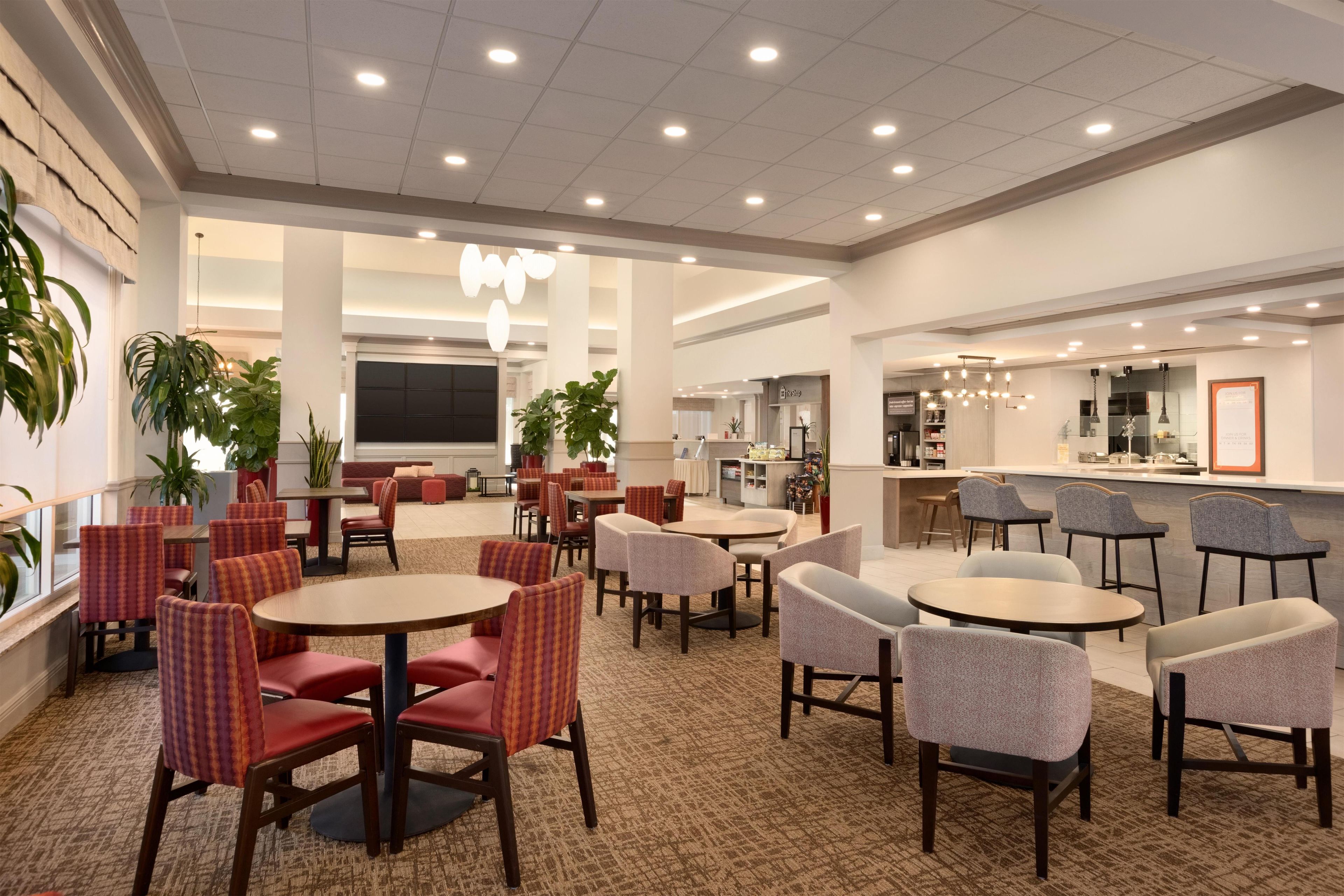 A hotel lobby with multiple seating areas, including round tables and chairs, a bar area, and a large screen.