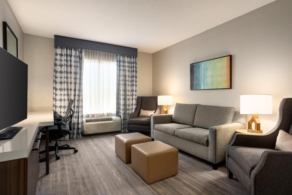 Een hotelkamer met een flatscreen-televisie, een bank, twee poefs, een bureau met stoel, een lamp en een schilderij aan de muur.