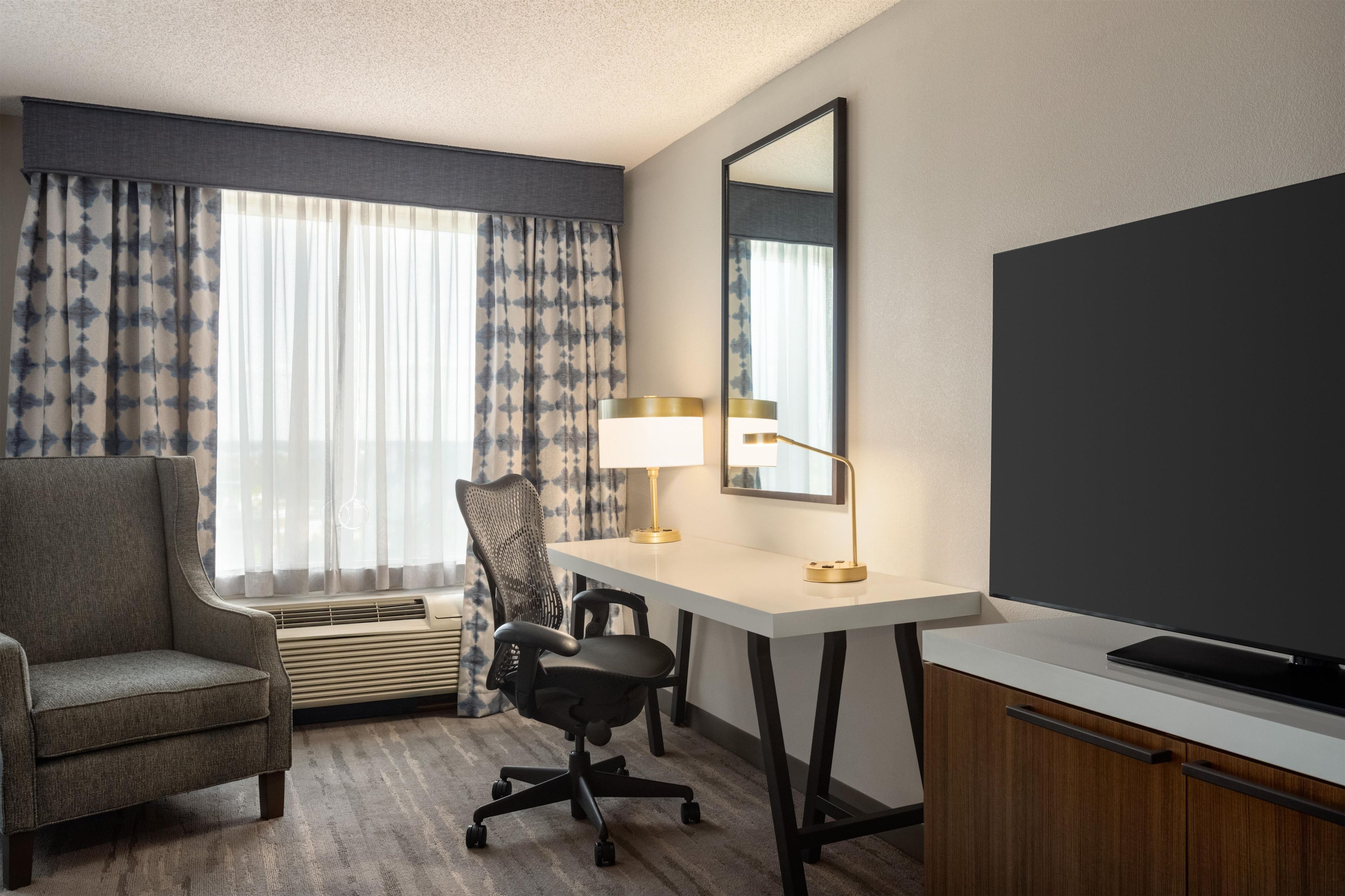 A hotel room with a desk, chair, air conditioning unit, and a television.